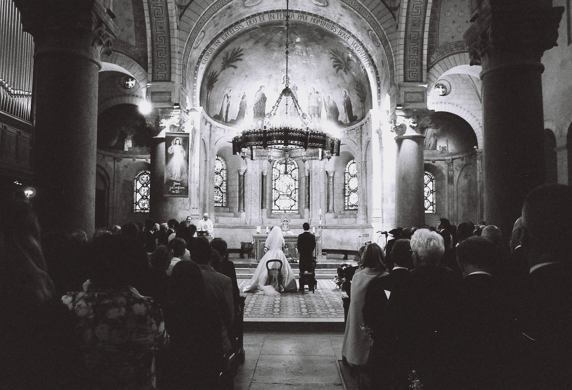Photographe corrèze. Deux mariés en costume traditionnel sont à l'autel d'une basilique Lyonnaise. Prise depuis le fond de l'église, la photo présente deux masses d'invités de chaque côté de l'allée centrale, guidant le regard vers les mariés debout au milieu du transept. Devant et au dessus d'eux, le décors grandiose de la basilique se dévoile en tons clairs illuminant la scène. Le grain de la photo argentique, en noir et blanc, est perceptible.